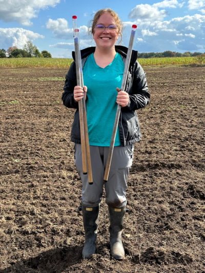 Graduate student Katelyn Anderson stands in a farm field, smiling and displaying sediment cores to the camera.